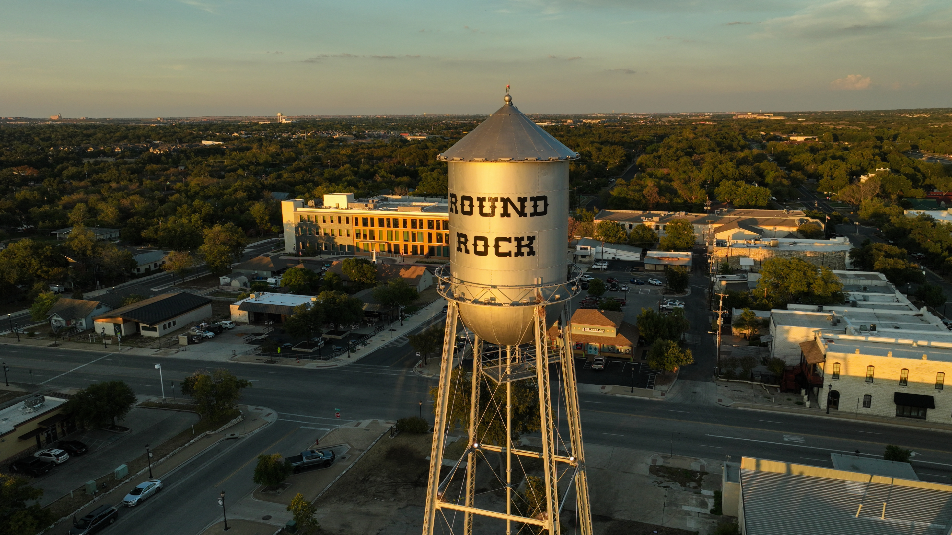 Aerial view of the Round Rock water tower in Texas, overlooking the downtown area with buildings, streets, and lush greenery during sunset.