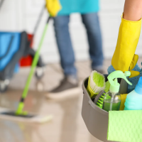 A person wearing yellow gloves and a teal cleaning apron is holding a cleaning bucket with various cleaning supplies while mopping a hardwood floor in a home. The image illustrates professional cleaning services with focus on hygiene and efficiency.