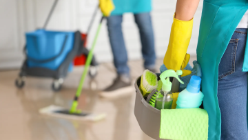 A person wearing yellow gloves and a teal cleaning apron is holding a cleaning bucket with various cleaning supplies while mopping a hardwood floor in a home. The image illustrates professional cleaning services with focus on hygiene and efficiency.