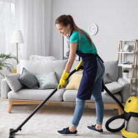 A woman in yellow gloves and an apron is vacuuming the carpet in a modern living room. The room features a comfortable couch, houseplants, and neutral-colored décor, showing a clean and organized space as part of a professional cleaning service.