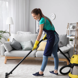 A woman in yellow gloves and an apron is vacuuming the carpet in a modern living room. The room features a comfortable couch, houseplants, and neutral-colored décor, showing a clean and organized space as part of a professional cleaning service.