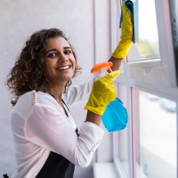 A woman in yellow gloves is smiling while cleaning a window with a spray bottle and cloth. This image represents our affordable cleaning service in Round Rock, TX, focusing on quality and customer satisfaction for all your home or office cleaning needs.